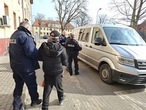 policjant prowadzi w kierunku nieoznakowanego radiowozu mężczyznę, który ma ręce skute kajdankami. Przed radiowozem stoją dwaj inni policjanci z innym zatrzymanym mężczyzną