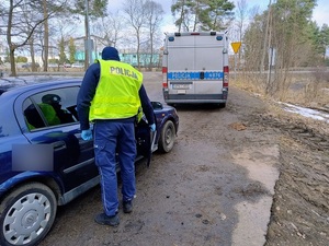 na piaszczystej drodze przy granatowym osobowym oplu stoi policjant ruchu drogowego. Policjant trzyma otwarte drzwi od strony pasażera tego samochodu. Przed oplem stoi oznakowany policyjny radiowóz