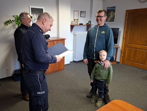 w gabinecie Komendanta Powiatowego Policji w Lęborku komendant odczytuje podziękowania kierowane do mężczyzny stojącego naprzeciw niego. Przy mężczyźnie stoi kilkuletni chłopiec. Mężczyzna się uśmiecha. Obok komendanta stoi jego zastępca