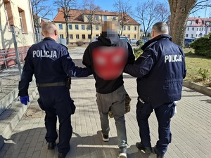 dwaj policjanci prowadzą zatrzymanego mężczyznę w kierunku nieoznakowanego radiowozu