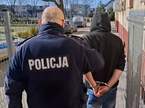 na zewnątrz budynku komendy policjant prowadzi mężczyznę, który ma ręce skute za plecami kajdankami