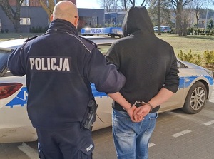 policjant prowadzi w kierunku oznakowanego radiowozu mężczyznę, który ma ręce skute za plecami kajdankami