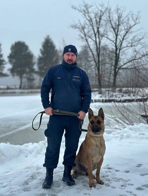 zima. Na ośnieżonym terenie stoi policjant, który trzyma na smyczy siedzącego obok psa służbowego