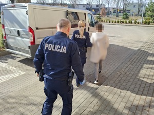 policjantka prowadzi zatrzymaną kobietę w kierunku nieoznakowanego radiowozu. Za nią idzie policjant