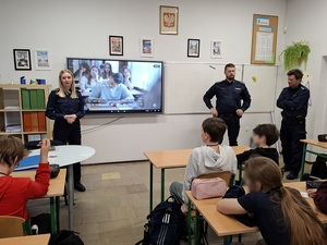 klasa szkolna. Przed siedzącymi w ławkach uczniami stoją: policjantka i dwaj policjanci. Za nimi na ekranie jest wyświetlony film profilaktyczny