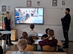 klasa szkolna. Przed siedzącymi w ławkach uczniami stoją: policjantka i policjant. Za nimi na ekranie jest wyświetlony film profilaktyczny