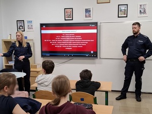 klasa szkolna. Przed siedzącymi w ławkach uczniami stoją: policjantka i policjant. Za nimi na ekranie jest wyświetlony informacja o instytucjach pomocowych