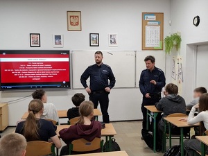 klasa szkolna. Przed siedzącymi w ławkach uczniami stoją dwaj policjanci. Za nimi na ekranie jest wyświetlona informacja o instytucjach pomocowych