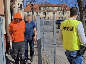 nieumundurowany policjant prowadzi mężczyznę, który ma ręce skute za plecami kajdankami. Przed nimi idzie drugi policjant, ubrany w kamizelkę odblaskową z napisem Policja