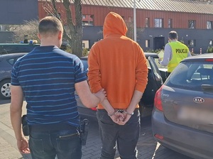 przed nieoznakowanym radiowozem stoi mężczyzna, który ma ręce skute za plecami kajdankami. Obok stoją dwaj policjanci