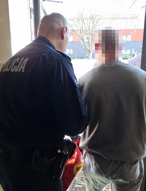 policjant wyprowadza z budynku komendy mężczyznę, który ma ręce skute kajdankami