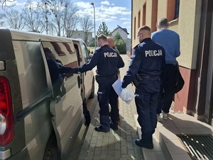 Przed nieoznakowanym radiowozem znajdują się dwaj policjanci i dwaj zatrzymani mężczyźni. Policjant z zatrzymanym stoją przed otwartymi drzwiami radiowozu, drugi policjant idzie z zatrzymanym w ich kierunku