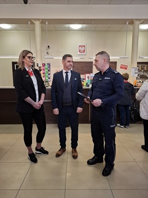 na poczcie w miejscu obsługi klientów stoją obok siebie komendant policji, pracownik i pracownica poczty. Komendant trzyma dyplom