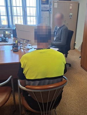 przy biurku przed komputerem siedzi policjant. Obok, przed biurkiem na krześle siedzi zatrzymany mężczyzna