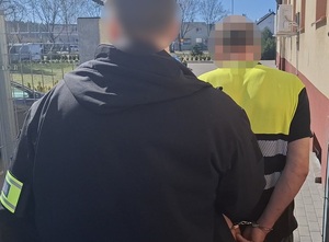 na zewnątrz komendy policjant kryminalny prowadzi mężczyznę, który ma ręce skute za plecami kajdankami