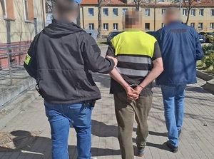 na zewnątrz komendy policjant kryminalny prowadzi mężczyznę, który ma ręce skute za plecami kajdankami. Obok idzie inny nieumundurowany policjant