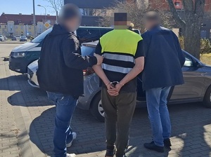 Przed nieoznakowanym radiowozem stoi mężczyzna, który ma ręce skute za plecami. Obok niego znajdują się dwaj nieumundurowani policjanci