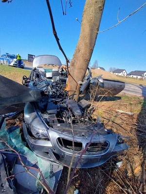 poza jezdnią na pasie zieleni znajduje się roztrzaskany o drzewo samochód osobowy. Na poboczu stoi policyjny radiowóz, a przy nim znajduje się policjant ruchu drogowego