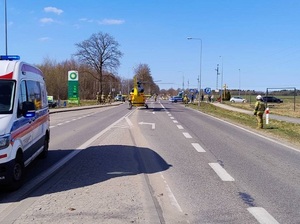 na środku jezdni ląduje śmigłowiec. Na jezdni stoi karetka pogotowia, przed śmigłowcem na drodze stoją policjanci i strażacy