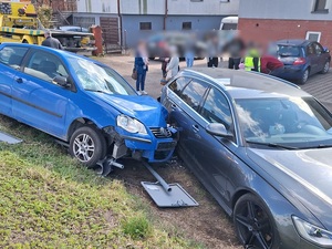 miejsce zdarzenia drogowego. Poza jezdnią na pasie zieleni niebieski Vw Polo wjechał w bok szarego osobowego samochodu. Pod VW leży złamany znak drogowy. Oba pojazdy mają wgniecioną karoserię. W Vw wystrzeliły poduszki powietrzne. Za tym autami stoją gapie