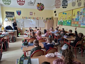 klasa szkolna. Przy biurku nauczyciela stoi policjantka. Na ekranie  tablicy wyświetla się film. W ławkach siedzą pierwszoklasiści