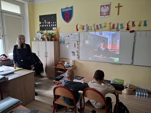 klasa szkolna. Przy biurku nauczyciela stoi policjantka. Na ekranie  tablicy wyświetla się film. W ławkach siedzą pierwszoklasiści
