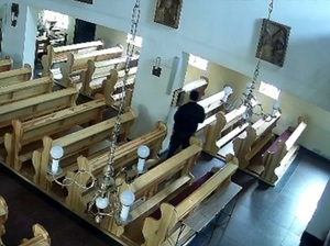 wnętrze kościoła. Przy ławkach na ścianie wisi skarbonka. Drzwiczki skarbonki są otwarte. Stoi przy niej mężczyzna ubrany na czarno, w czarnej czapce