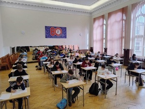 w szkolnej auli przy ławkach siedzą pojedynczo uczniowie, którzy rozwiązują test