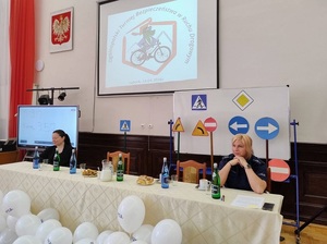 w szkolnej auli przy stole siedzi policjantka i nauczycielka. Za policjantką stoją ćwiczeniowe znaki drogowe. Na ekranie wyświetla się informacja o Turnieju Bezpieczeństwa Ruchu Drogowego