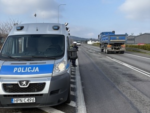 na poboczu drogi stoi oznakowany policyjny radiowóz. Przy nim stoi policjant ruchu drogowego, który mierzy prędkość przejeżdżających samochodów za pomocą ręcznego miernika