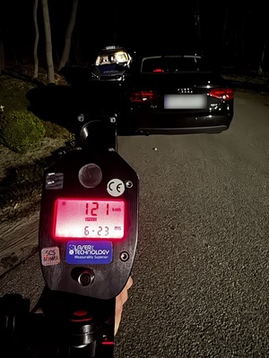 pora wieczorowa. Na poboczu stoi ciemne osobowe Audi. Za nim widać trzymany w ręce policjanta ręczny miernik prędkości, na wyświetlaczu którego widać zmierzoną prędkość 121 km/h