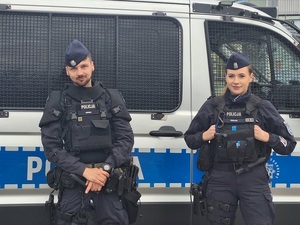 przy oznakowanym radiowozie typu "bus' stoi dwoje policjantów służby patrolowej: uśmiechnięci policjant i policjantka