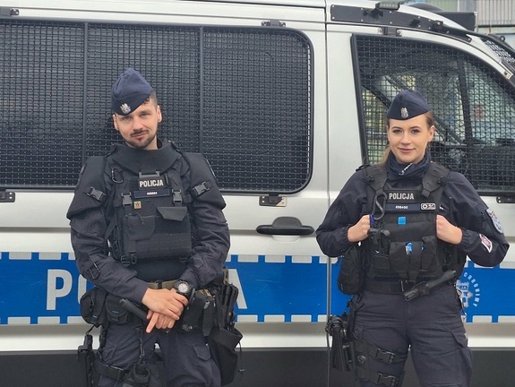 przy oznakowanym radiowozie typu "bus' stoi dwoje policjantów służby patrolowej: uśmiechnięci policjant i policjantka