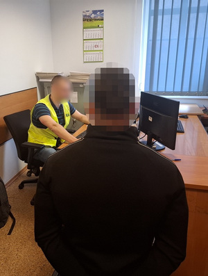 w pokoju przesłuchań obok biurka siedzi na krześle zatrzymany mężczyzna. Przy biurku przed komputerem siedzi nieumundurowany policjant w kamizelce odblaskowej z napisem "Policja"