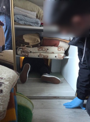 przez mały otwór w szafie pod dolną półką przeczołguje się mężczyzna. Przed szafą i obok niej znajdują się policjanci kryminalni.