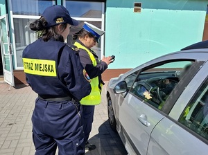 w strefie zamieszkania na targowisku miejskim przy osobowym samochodzie stoją policjantka ruchu drogowego i strażniczka miejska. Policjantka sprawdza auto w systemach policyjnych