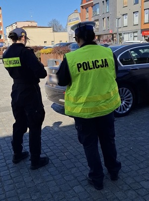 w centrum miasta przy osobowym samochodzie stoją policjantka ruchu drogowego i strażniczka miejska