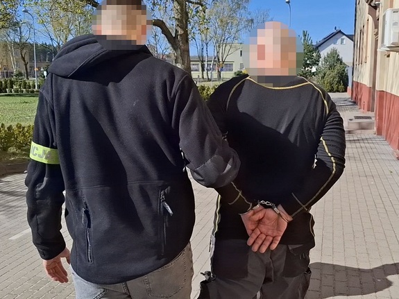 na zewnątrz komendy Policji funkcjonariusz kryminalny prowadzi mężczyznę, który ma ręce skute za plecami kajdankami