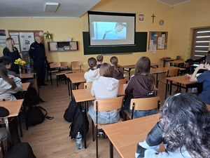 w szkolnej klasie, przed siedzącymi w ławkach uczniami stoją policjantka i policjant. Uczniowie oglądają wyświetlony na tablicy film profilaktyczny