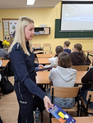 w szkolnej klasie policjantka rozdaje siedzącym w ławkach uczniom ulotki profilaktyczne