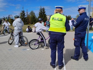 na placu znajdują się dzieci na rowerach. Obok stoją policjant i policjantka ruchu drogowego