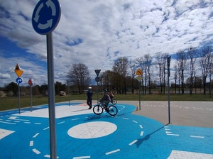 po miasteczku rowerowym jedzie rowerami dwoje uczniów. Obok stoi policjant ruchu drogowego, który obserwuje ich jazdę