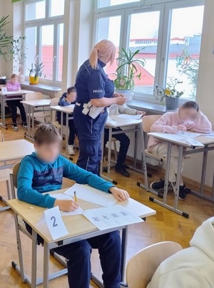 w klasie szkolnej uczniowie rozwiązują test. Pomiędzy ławkami chodzi policjantka ruchu drogowego