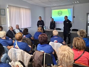 w auli komendy przed siedzącymi w rzędach seniorami stoi troje policjantów. Za nimi na ekranie wyświetla się prezentacja multimedialna
