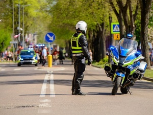 na ulicy obok oznakowanego policyjnego motocykla stoi policjant ruchu drogowego. Przed nim widać radiowóz, a za radiowozem uczestników przemarszu