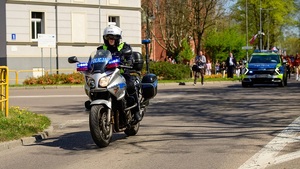 ulicą jedzie motocyklem policjant ruchu drogowego. Za nim widać radiowóz, a za radiowozem uczestników przemarszu