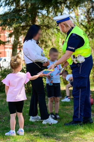 policjantka ruchu drogowego przekazuje stojącej obok dorosłej opiekunki dziewczynce książeczkę profilaktyczną