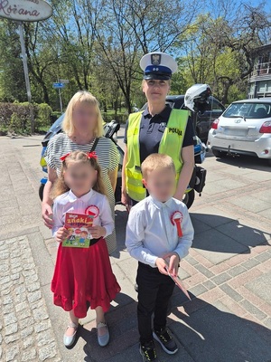 na chodniku stoi policjantka ruchu drogowego w towarzystwie kobiety i dwojga dzieci. Dzieci trzymają książeczki profilaktyczne o znakach drogowych