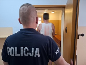 w celi stoi zatrzymany mężczyzna. Za nim przy drzwiach stoi policjant służby dyżurnej