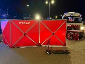 pora wieczorowa. Na jezdni leży hulajnoga elektryczna. Przed nią jest rozstawiony czerwony parawan straży pożarnej. Obok stoi karetka